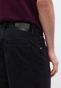 Juodi džinsai iš džinso audinio su odiniu Calvin Klein logotipo dirželiu, pasižymintys klasikiniu penkių kišenių dizainu ir subtiliais siuvimo elementais.