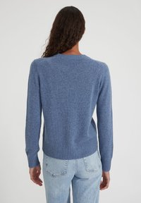 Femme aux longs cheveux bouclés portant un pull en maille bleu et un jean clair, de dos contre un fond uni clair.