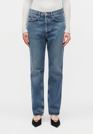 Άτομο που φοράει μπλε τζιν με ευθεία γραμμή και ψηλή μέση, λευκό μπλουζάκι με μακριά μανίκια και μαύρα παπούτσια με μυτερή μύτη σε λευκό φόντο.