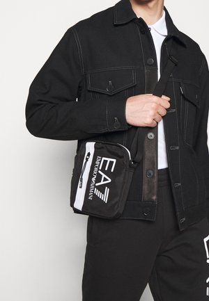 Zwarte stoffen schoudertas met witte accenten en logo. Compacte rechthoekige vorm met ritssluiting en verstelbare band.