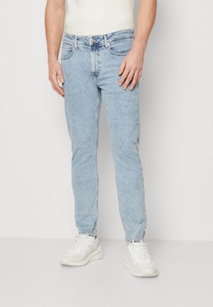Mann trägt hellblaue Slim-Fit-Jeans und weiße Diesel-Sneaker, steht vor einem schlichten weißen Hintergrund.