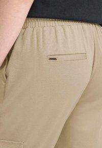 Beige joggebukser med elastisk liv, en enkelt baklomme med horisontal åpning og liten sølvdetalj. Jevn tekstur.