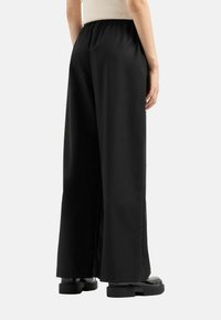 Pantaloni neri a gamba larga con elastico in vita, texture liscia e vestibilità rilassata. Indossati con scarpe con la zeppa nere e voluminose.