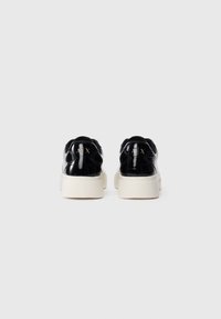 Baskets en cuir verni noir avec des semelles épaisses blanches, vues de l'arrière sur un fond blanc, arborant un logo doré "A|X" sur chaque talon.