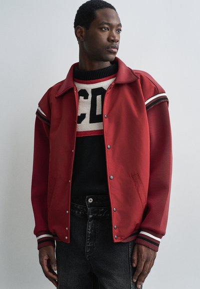 Uomo che indossa una giacca varsity rossa sopra un maglione bianco e nero con lettere in grassetto, in piedi davanti a uno sfondo grigio chiaro uniforme, guardando verso destra.