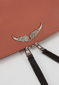 Torba ze skórki w kolorze brzoskwiniowym z metalowym akcentem w kształcie skrzydła w kolorze srebrnym, podwójnymi czarnymi rączkami i metalowym zamkiem błyskawicznym z kwadratowymi wycięciami.