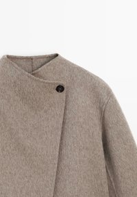 Cappotto di lana beige con superficie testurizzata, dotato di chiusura diagonale e bottone nero. Linee pulite e design minimalista.