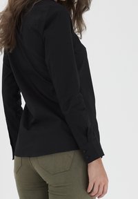 Black long-sleeve shirt with a structured design, buttoned cuffs, worn with olive green fitted pants, showing smooth fabric and tailored fit.