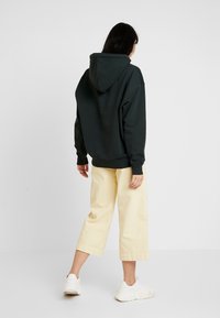 Tmavě zelená oversize mikina s centrálním švem na zádech a přední kapsou ve stylu klokan, spárovaná se světle žlutými širokými zkrácenými kalhotami.