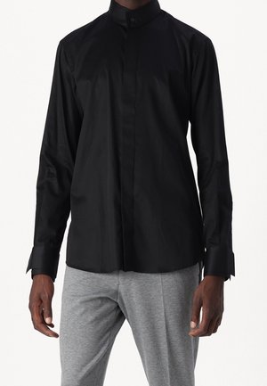 Czarna koszula z długim rękawem z ukrytymi przednimi guzikami i wydłużonymi mankietami, noszona z jasnoszarymi spodniami przez stojącą osobę na jednolitym tle.