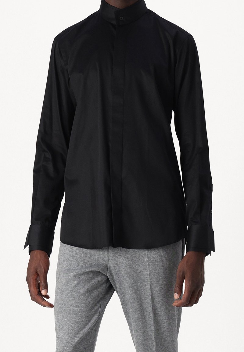 Czarna koszula z długim rękawem z ukrytymi przednimi guzikami i wydłużonymi mankietami, noszona z jasnoszarymi spodniami przez stojącą osobę na jednolitym tle.