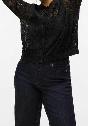 Vrouw draagt een zwarte kanten blouse met knopen en donkerblauwe jeans met hoge taille, armen boven het hoofd geheven tegen een witte achtergrond.