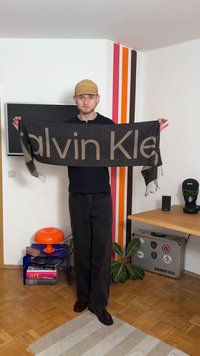 Écharpe noire avec logo "Calvin Klein" marron, motif texturé, bords à franges, tenue dans une pièce bien éclairée avec un sol en bois.