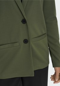 Oliwkowy żakiet o gładkiej fakturze, z dwoma czarnymi guzikami i łukowatymi kieszeniami z przodu; długie rękawy o prostym kroju.