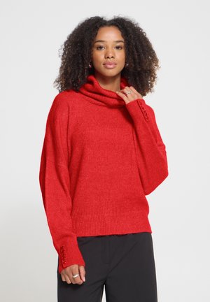 Femme aux cheveux bouclés portant un pull à col roulé rouge avec des poignets boutonnés et un pantalon noir sur un fond uni.