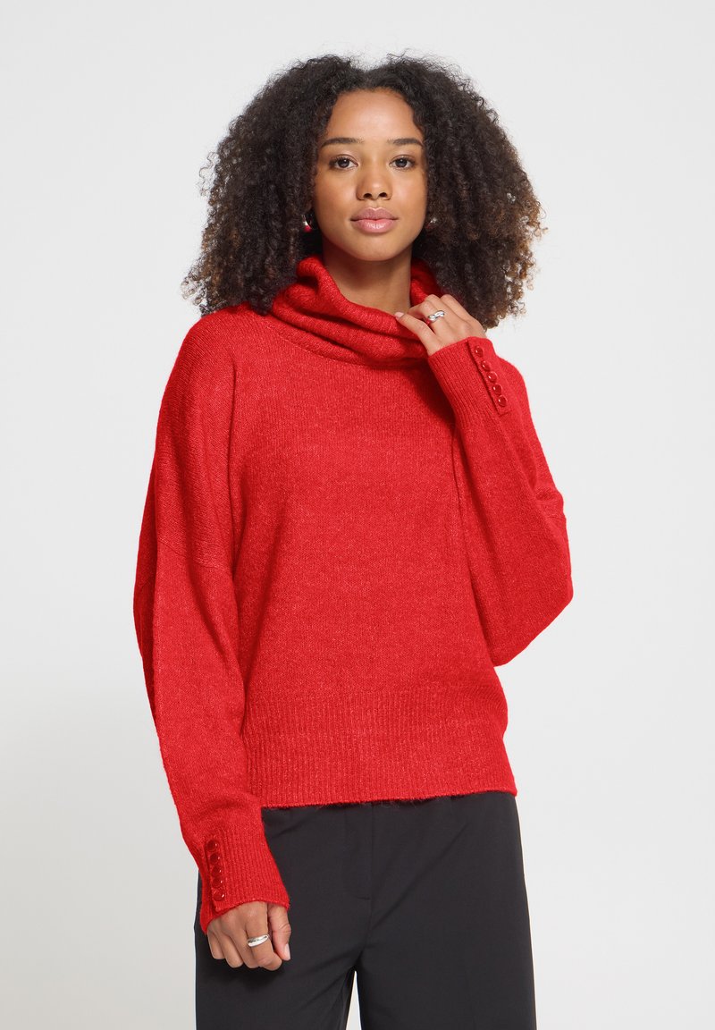 Femme aux cheveux bouclés portant un pull à col roulé rouge avec des poignets boutonnés et un pantalon noir sur un fond uni.