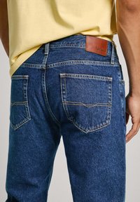 Jeans de mezclilla en azul oscuro con una textura ligeramente desgastada, que cuentan con dos bolsillos traseros y una etiqueta de cuero marrón en la cinturilla.