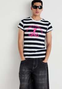Tânăr purtând ochelari de soare negri, un tricou alb și negru cu dungi și text roz "Sunt petrecerea de după", și pantaloni largi cu un model închis.