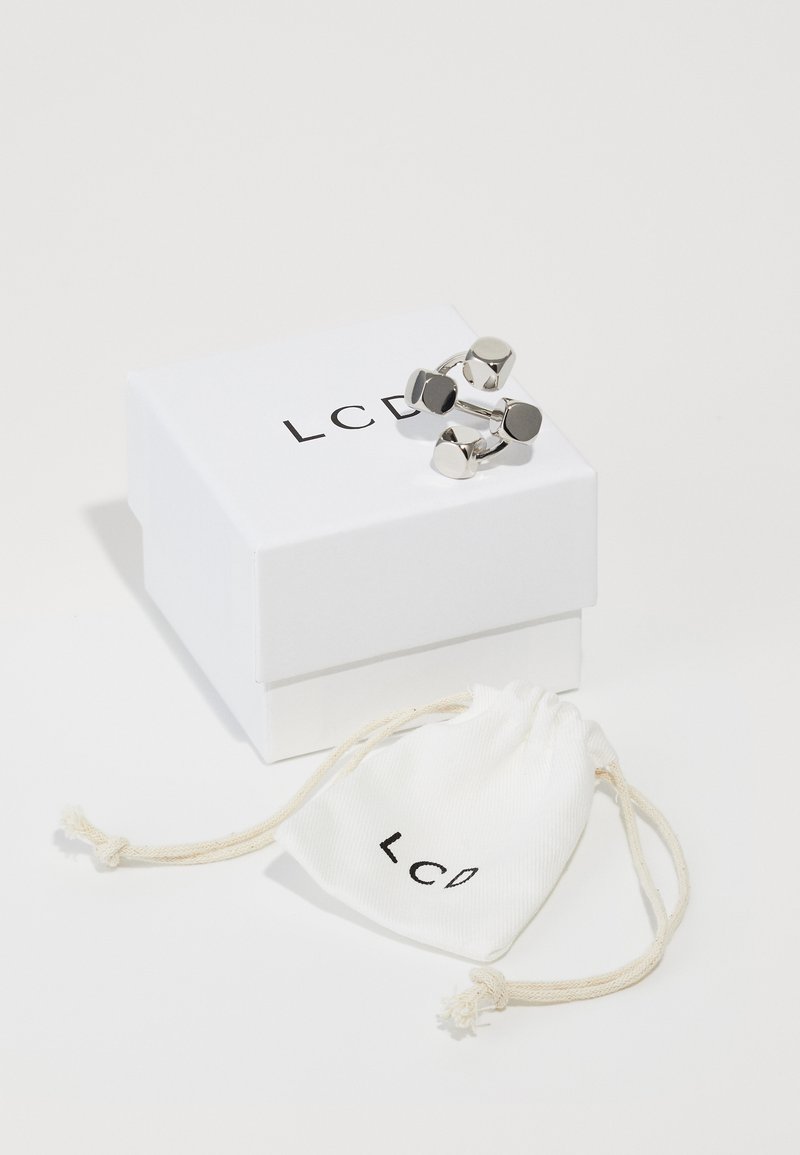 Gemelli da polso color argento con forme geometriche, sistemati su una scatola bianca con il testo 'LCD', accanto a una borsa crema con il logo 'LCD'.