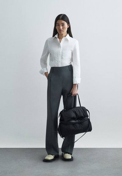 Λευκό πουκάμισο με γιακά, γκρι παντελόνι ψηλής μέσης, μαύρη τσάντα με μαλακή υφή, παπούτσια κρέμα και μαύρα, ποζάροντας σε ουδέτερο φόντο.