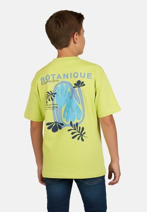 Jongen draagt een limoengroene T-shirt met een blauw en zwart botanisch ontwerp en de tekst "BOTANIQUE" op de achterkant, gecombineerd met een blauwe spijkerbroek.