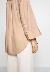 Beige pitkähihaine paita, jossa on kaareva helma ja napitettu hihansuissa, valmistettu sileästä kankaasta. Yhdistetty valkoisiin leveälahkeisiin housuihin.