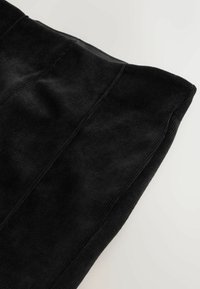 Mustat korkeavyötäröiset corduroy-housut näkyvillä saumoilla, asetettuna tasaiselle valkoiselle pinnalle, näyttäen kankaan tekstuuria ja ompelutietoja.