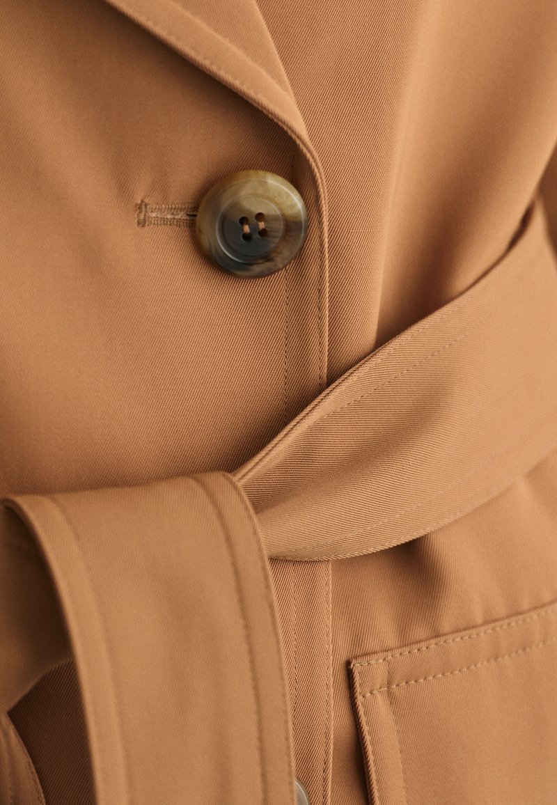 Blazer beige avec un grand bouton, une ceinture apparente, et un tissu lisse avec une texture subtile. Comprend une poche avant avec des accents de couture.