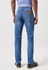 Vaqueros de mezclilla azul con corte recto, que presentan un parche de cuero en la parte trasera, dos bolsillos traseros y una textura sutil. Combinados con zapatos negros.