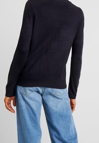 Persona che indossa un maglione nero a maniche lunghe aderente e jeans di denim blu chiaro, vista da dietro su uno sfondo semplice.