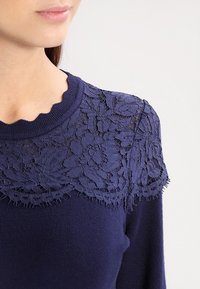 Mujer vistiendo un suéter azul marino con un yugo de encaje floral y escote con volantes, contra un fondo claro y sencillo.