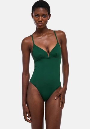 Femme aux cheveux courts portant un maillot une-pièce vert foncé à fines bretelles et décolleté en V profond, debout devant un fond gris uni.