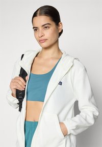 Witte fleece hoodie met een capuchon en voorzakken, gedragen boven een teal crop top. Het model houdt een zwarte rugzakriem vast.