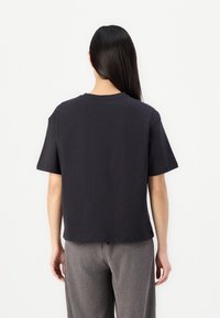 Femme aux longs cheveux noirs portant un t-shirt noir uni à manches courtes et un pantalon gris, debout de dos face au spectateur.