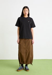 Svart bomulls t-skjorte med en liten grafisk detalj, kombinert med et brunt skjørt med elastisk midje. Modellen har på seg grå joggesko med blå snørebånd.