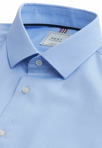 Svijetloplava slim-fit košulja od teksturirane tkanine. Ima klasičan ovratnik, dva bijela gumba i oznaku marke s šarenim detaljima.