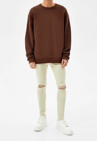 Sudadera marrón con hombros caídos, combinada con jeans ajustados rotos color crema y zapatillas blancas con acentos dorados.