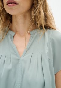 Femme aux cheveux clairs et ondulés portant un chemisier plissé vert clair avec un col en V, montrée du cou jusqu'à la partie supérieure de la poitrine.