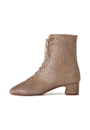 Donna in piedi al chiuso sorridente, indossa una maglia a coste azzurro chiaro a maniche lunghe, pantaloni larghi scuri, una cintura con motivo e stivaletti beige con lacci.