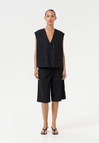 Črn laneni brezrokavnik z globokim V-izrezom, dvema sprednjima žepoma in gumbnim zapiranjem, v kombinaciji z ujemajočimi se koleni dolgimi culottes.