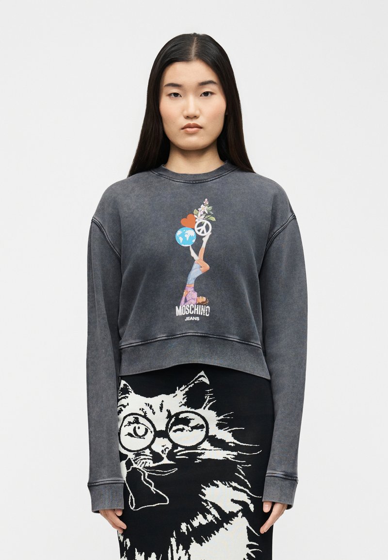 Νεαρή γυναίκα που φοράει ένα ξεθωριασμένο μαύρο φούτερ με πολύχρωμο σχέδιο και μαύρη φούστα με μεγάλο λευκό γάτο με γυαλιά.