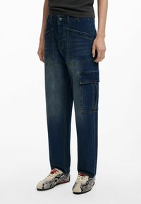 Pantalones cargo de mezclilla anchos y de color azul oscuro, con múltiples bolsillos frontales y un bolsillo lateral, que cuentan con un cierre de botón clásico y una textura desgastada.