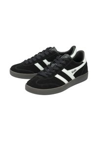 Svarta mockaskor med vita läderdetaljer och tre vita ränder. Platt gummisula och snörningsdesign. Logotyp på sidan.