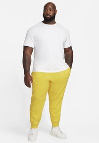 Camiseta blanca de manga corta, pantalones de chándal amarillos y zapatillas blancas. Los pantalones de chándal tienen puños elásticos y un pequeño logotipo en el lateral. Textura suave en toda la prenda.