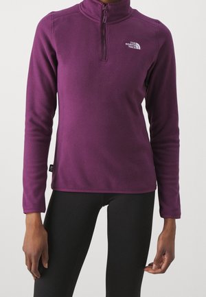 Kobieta nosząca fioletowy polar z suwakiem do ćwiartki i logo "The North Face", połączony z czarnymi legginsami.