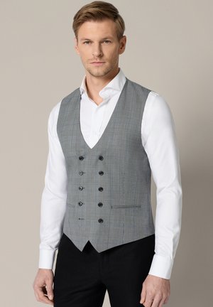 Mężczyzna w białej koszuli i szarej dwurzędowej kratkowanej kamizelce stoi na jednolitym tle.