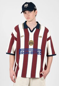 Jeune homme portant une casquette bleu marine et une chemise oversize rayée bordeaux et blanche avec le texte «ROHIBITED» ainsi qu'un pantalon beige sur un fond uni.