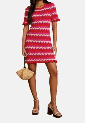Femme portant une mini robe en tricot zébrée rouge, rose et crème avec des manches courtes, tenant un sac à main en forme d'éventail tissé et portant des talons compensés beige.
