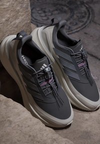 Ζευγάρι ασπρόμαυρα αθλητικά παπούτσια Adidas με χοντρές σόλες και ελαστικά κορδόνια τύπου bungee, τοποθετημένα σε πέτρινο πλακόστρωτο κοντά σε βράχια.