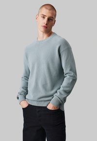  Lichtblauwe gebreide trui met een gestructureerd patroon, ronde hals, geribde manchetten en onderrand. Model draagt donkerblauwe jeans, met de handen in de zakken.